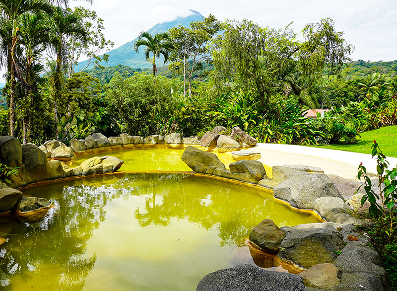 Arenal Volcano smoking crater with guests relaxing in natural hot springs surrounded by rainforest - Macaws Adventure Tours by Pablo C